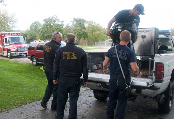 Four men loading a grill and a smoker into a pick up truck parked in a driveway. Some wear clothing labeled "FIRE". Red vehicles are parked in the background