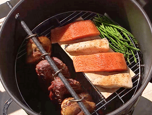 Hinged cooking grate in barrel smoker from above, lid off. Half of the hinged grate is dropped down to let ribs hang past the grate level. Fish and veggies are cooking on the other half.
